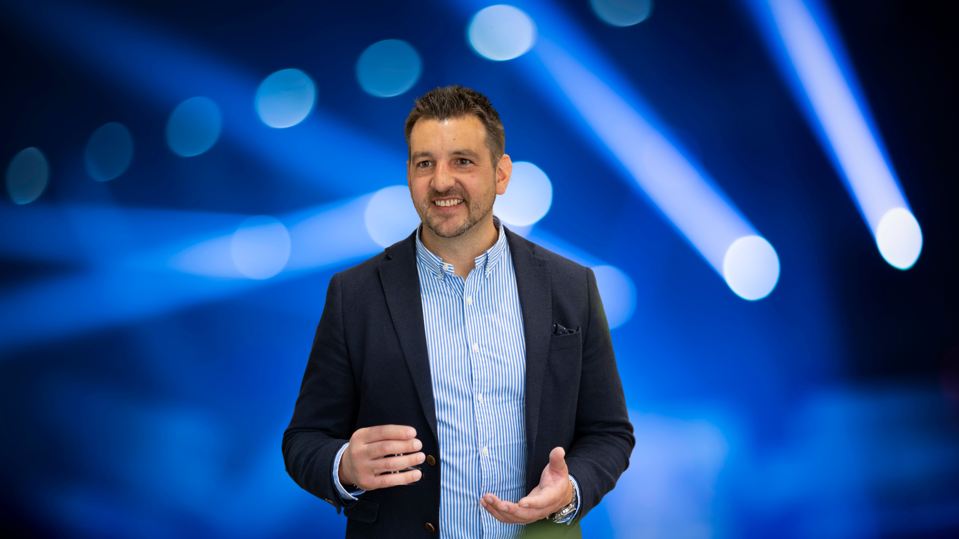 Man wearing a blazer and shirt, smiling and gesturing in front of a blue background with lights.