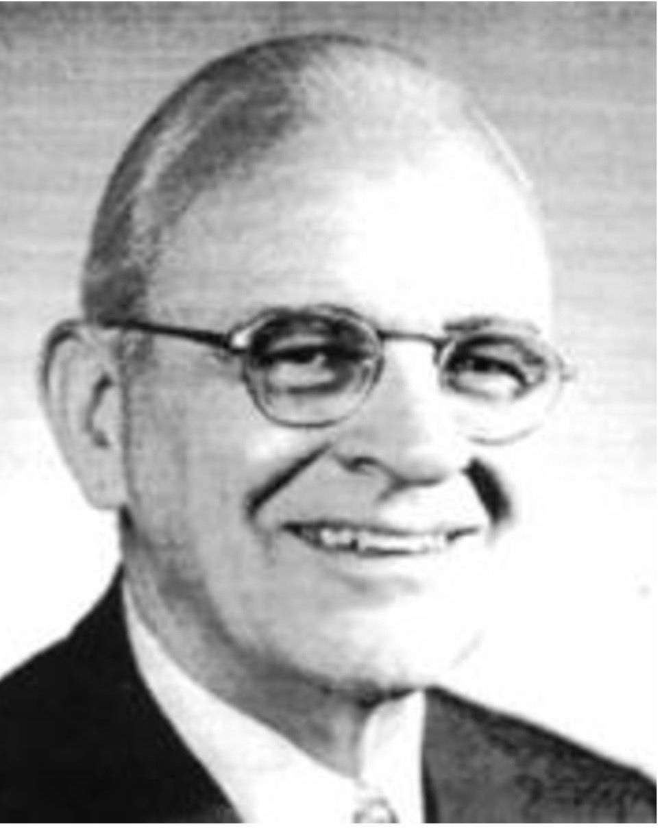 Black and white portrait of a man wearing glasses and a suit, smiling at the camera.