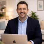Portrait of a smiling man in a navy blazer and light blue shirt seated at a desk with a laptop; cosy home office background with plant and framed art