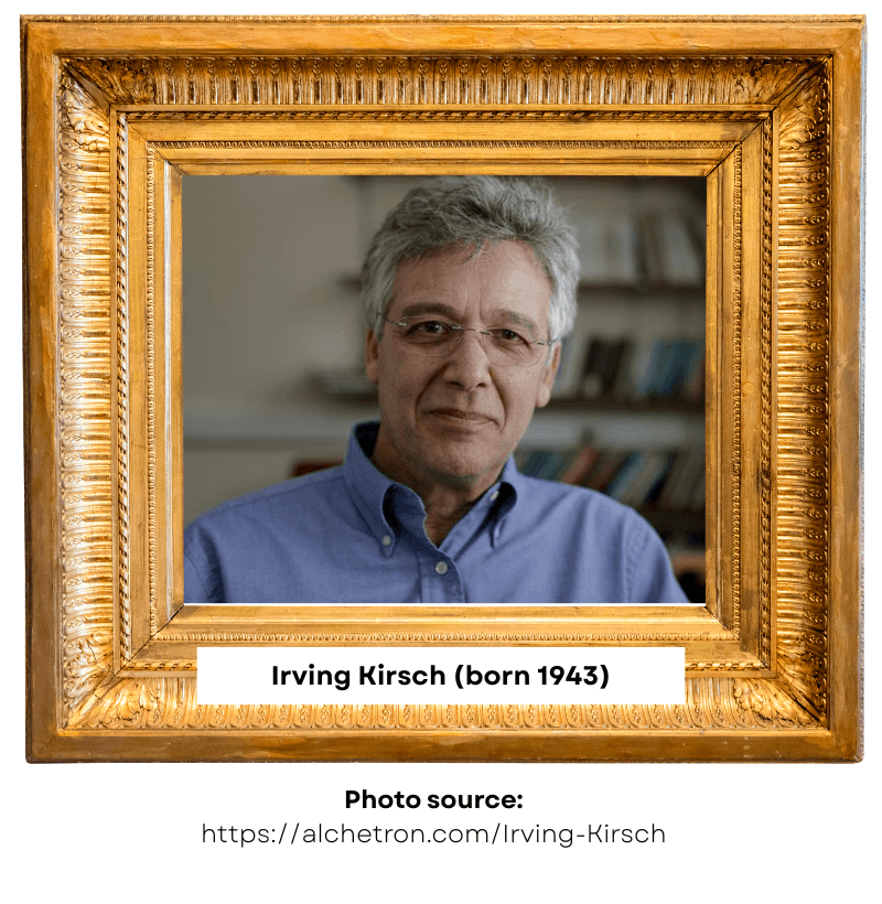 Irving Kirsch with grey hair and glasses, wearing a blue shirt, photographed in front of a soft-focus bookshelf, framed by an ornate gold frame