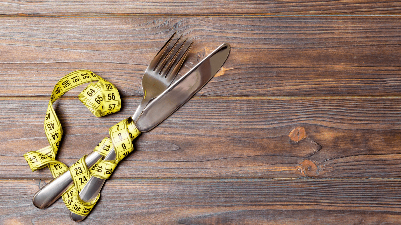 Close-up of a yellow measuring tape wrapped around a fork and knife on a rustic wooden surface, highlighting metal cutlery and the wood grain